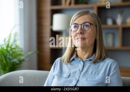 Une femme d'âge moyen réfléchie portant des lunettes et une chemise bleue est assise sur un canapé dans un environnement domestique confortable. Banque D'Images