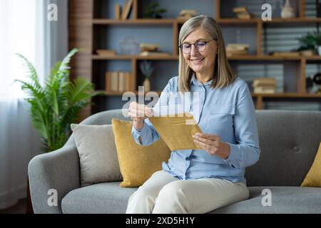 Vieille dame avec des lunettes assis sur un canapé, lisant joyeusement une lettre dans son salon confortable. Banque D'Images