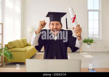 Homme dans la robe de gradué assis à la table avec diplôme faisant le geste oui pendant l'appel vidéo en ligne. Banque D'Images