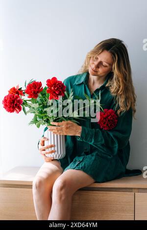 Femme en robe verte est assise sur un banc en bois, tenant un vase blanc rempli de pivoines de fleurs rouges vibrantes, sur fond blanc. Banque D'Images