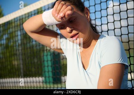 Femme fatiguée prenant une pause de jouer au tennis, essuyant la sueur du front. Montre de la détermination, de la fatigue et de l'effort dans les sports de tennis. Extérieur ensoleillé s Banque D'Images