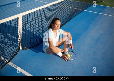 Une femme en tenue sportive fait une pause sur un court de tennis, assise contre le filet, tenant une raquette et une balle. La scène transmet la détente et la dissuasion Banque D'Images