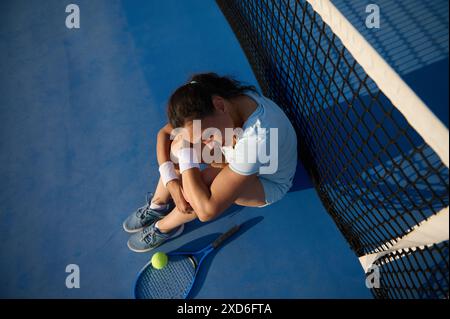 Vue aérienne d'une joueuse de tennis fatiguée assise sur un court bleu, appuyée contre le filet, avec une raquette et une balle à ses côtés. Banque D'Images