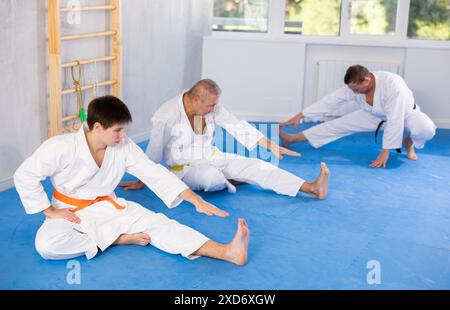 Hommes d'âges différents dans un kimono blanc faisant des étirements musculaires avant l'entraînement de karaté ou de judo Banque D'Images