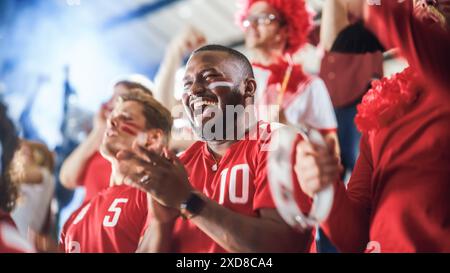 Sport Stadium Soccer match : une foule diversifiée de fans encouragent leur équipe rouge à gagner. Les gens célèbrent l'attribution d'un but, la victoire de championnat. Groupez les gens avec des visages peints acclamer, crier, s'amuser Banque D'Images