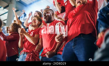 Sport Stadium Soccer match : une foule diversifiée de fans encouragent leur équipe rouge à gagner. Les gens célèbrent l'attribution d'un but, la victoire de championnat. Groupez les gens avec des visages peints acclamer, crier, s'amuser Banque D'Images