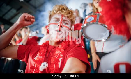 Stadium Sport Event : selfie Portrait de l'homme avec le visage peint se tenant et se tirant sur smartphone, encouragements pour l'équipe rouge pour gagner, hurler, célébrer la victoire du championnat avec la foule Banque D'Images