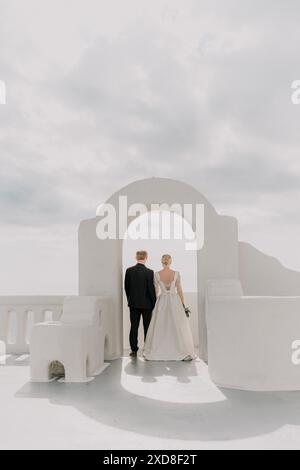 Une mariée et le marié marchent à travers une arche blanche par une journée ensoleillée. La mariée porte une robe blanche et le marié porte un costume. L'arche Banque D'Images