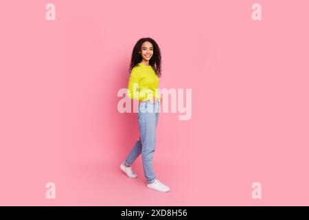 Photo pleine longueur du corps de la jeune fille avec les cheveux brune ondulés posant dans un pull vert de tenue décontractée et un Jean denim isolé sur fond de couleur rose Banque D'Images