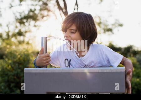 Un jeune garçon boit de l'eau d'une fontaine de parc par une belle journée ensoleillée. Il porte un t-shirt blanc et a les cheveux bruns. Banque D'Images