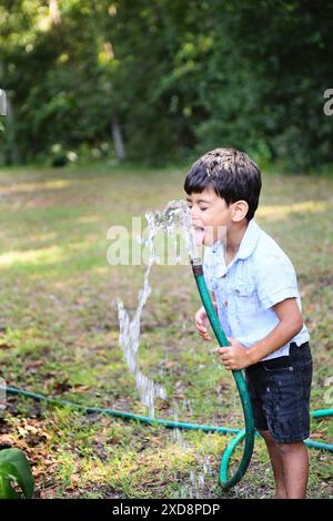 Un jeune garçon buvant de l'eau d'un tuyau d'arrosage dans une cour arrière. Banque D'Images