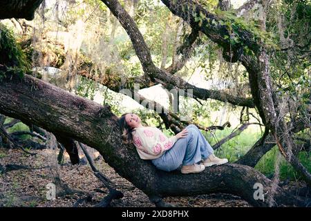 Jeune femme à la mode aux cheveux blonds repose sur un grand tronc d'arbre Banque D'Images
