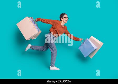 Photo du corps entier de la femme senior attrayante exécutant des sacs à provisions habillés élégants vêtements bruns isolés sur fond de couleur cyan Banque D'Images