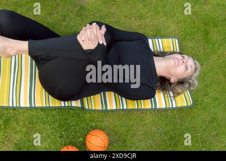 Une femme en survêtement noir sur l'herbe faisant de la gymnastique à la maison dans le jardin. Mains bloquées sur les genoux Banque D'Images