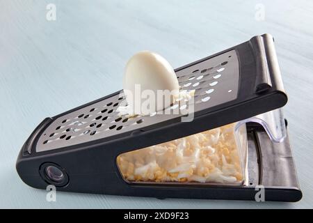 Oeuf de poule bouilli râpé sur une râpe en métal avec un récipient sur une table en bois bleu. Banque D'Images