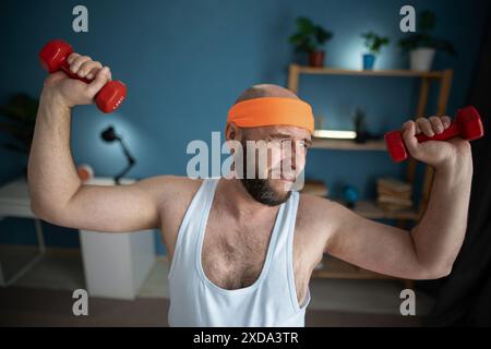 Homme d'âge moyen soulevant des haltères à la maison, luttant avec l'exercice, portant un bandeau orange et un débardeur blanc, fond bleu Banque D'Images