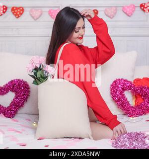 Jeune modèle brune sur le lit décoré pour les vacances romantiques de la Saint-Valentin. Haut Uni. Décorations de coeur et de rose autour d'elle. Banque D'Images