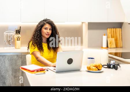 Beauté brune femme latine jeune créateur de contenu travaillant à la maison à l'aide d'un ordinateur portable tout en prenant le petit déjeuner le matin Banque D'Images