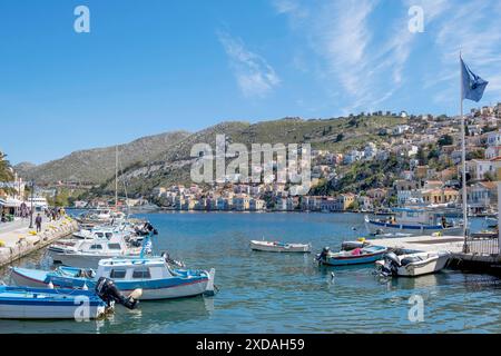 Un port tranquille avec plusieurs bateaux dans l'eau, flanqué des deux côtés par des étendues de terre vallonnées avec des maisons dispersées, sous un ciel bleu clair, Symi Banque D'Images