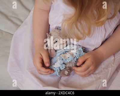 Agneau en peluche fabriqué à la main dans les mains de Blonde Toddler Girl contre une robe de coton rose pastel en toile de fond. Gros plan horizontal. Banque D'Images