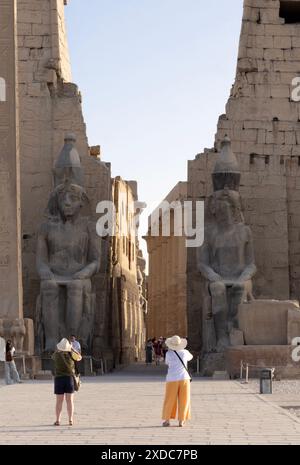 Deux femmes touristes chapeautées photographiant le temple de Louxor et les deux statues colossales de Ramsès II sur les rives est du Nil, en Égypte. Banque D'Images