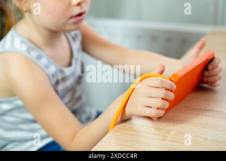 Jeune fille, enfant d'âge préscolaire assis pendu au-dessus de la table, engrossé dans le jeu de téléphone pendant une période prolongée, effets négatifs avec le cou coudé et les épaules roun Banque D'Images