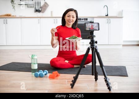 Femme en vêtements d'activité mangeant de la salade tout en enregistrant la vidéo de fitness dans la cuisine Banque D'Images