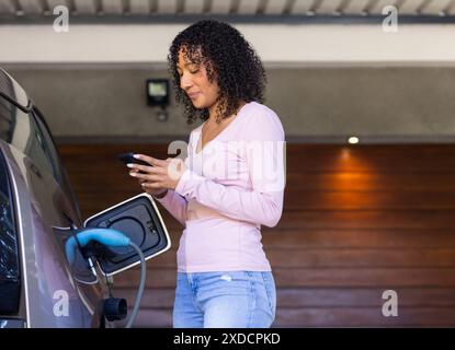 Femme chargeant la voiture électrique, en utilisant le smartphone dans le garage à la maison Banque D'Images