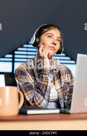 Port d'écouteurs et chemise à carreaux, jeune femme assise au bureau avec ordinateur portable et tasse Banque D'Images