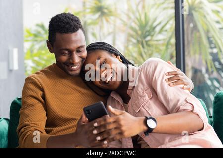 Assis sur le canapé, jeune couple souriant tout en regardant le smartphone ensemble Banque D'Images