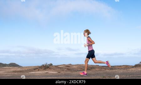 Une femme athlétique mince court sur un chemin de terre avec une chemise bleue et un short rose. Le ciel est clair et bleu, et il y a quelques nuages dans le ciel Banque D'Images