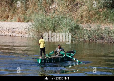 Un homme égyptien vêtu de façon traditionnelle rentre tandis qu'un garçon vêtu d'une robe moderne court un filet de pêche vers le bas du bateau vert dans les eaux du Nil. Banque D'Images