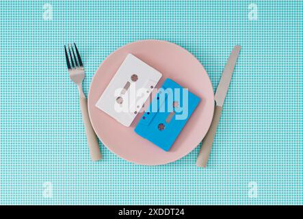 Cassettes audio sur plaque rose avec couteau et fourchette sur fond blanc et bleu. Nourriture minimale avec idée de musique rétro. Banque D'Images