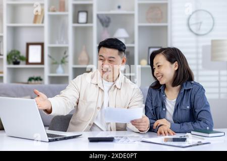 Jeune famille asiatique souriante, homme et femme assis à la maison à table devant un ordinateur portable et faisant le budget et les documents financiers. Banque D'Images