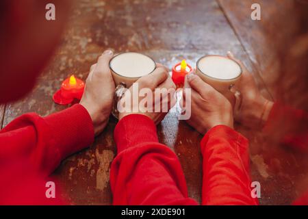 Gros plan d'un couple tenant des boissons chaudes dans les mains, avec une rangée de bougies en forme de cœur sur une table en bois devant eux. Le couple est w Banque D'Images