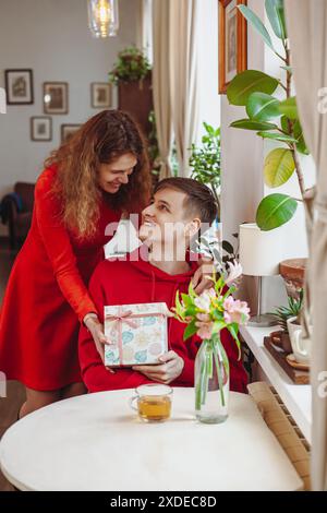 Une femme en robe rouge se tient derrière un homme assis à une table dans un café, lui donnant un cadeau emballé. L'homme sourit et tient le cadeau pendant que le w Banque D'Images