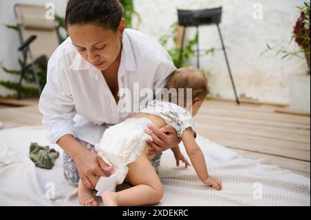 Une mère change soigneusement la couche de son bébé sur un patio extérieur, transmettant des soins et des liens dans un cadre naturel. La scène est intime et capture Banque D'Images