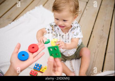 Tout-petit souriant jouant avec des jouets éducatifs colorés sur une terrasse en bois, s'engageant dans des activités d'intérieur amusantes et l'apprentissage des formes. Banque D'Images