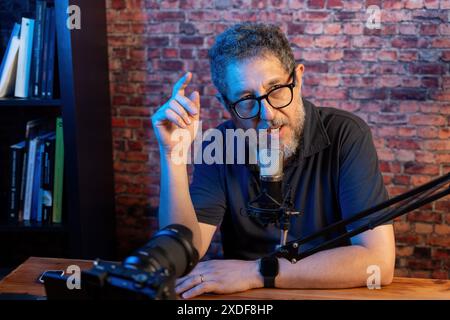 Un homme d'âge moyen avec des lunettes et une barbe podcasting avec un microphone, gestes et parler, fond de mur de briques, diffusion en ligne, contenu c Banque D'Images