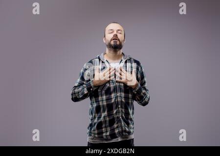 Homme barbu avec les yeux fermés tenant les deux mains sur sa poitrine, éprouvant de l'inconfort et de la douleur. Il se tient debout sur un fond gris Banque D'Images