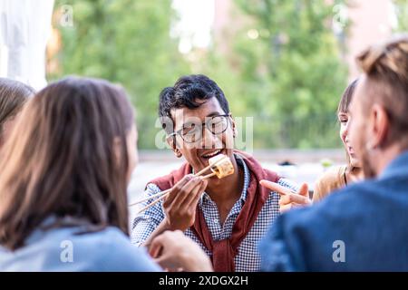 Amis appréciant un repas en plein air avec un homme sur le point de manger des sushis modernes. Ils rient et interagissent, créant une atmosphère vivante et sociale. Banque D'Images