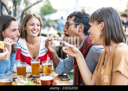 Groupe d'amis riant et appréciant la nourriture et les boissons à l'extérieur. Le groupe diversifié partage un repas amusant et animé, avec une personne mangeant de manière ludique Banque D'Images