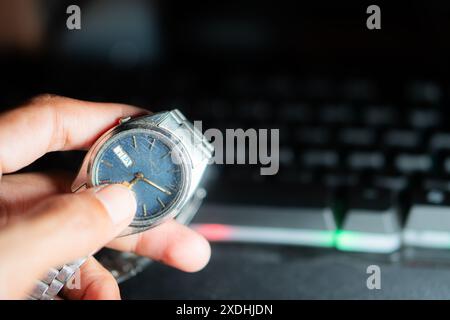 Gros plan d'une personne ajustant l'heure sur une montre-bracelet avec un cadran bleu, devant un clavier d'ordinateur. Banque D'Images