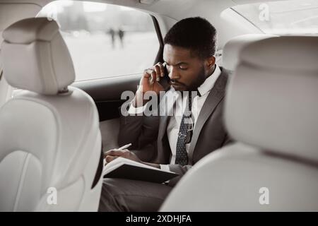 Homme d'affaires noir prenant des notes sur l'appel téléphonique dans le siège arrière de voiture Banque D'Images