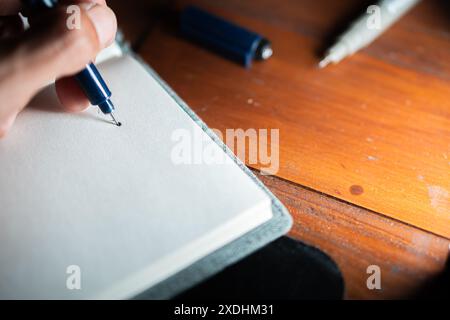 Gros plan d'une personne écrivant sur un cahier vierge avec un stylo. Le cahier est placé sur une table en bois avec d'autres articles de papeterie autour. Banque D'Images