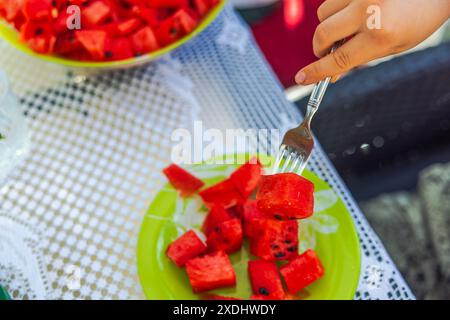 Vue rapprochée de la main de l'enfant avec la fourchette ramassant le morceau tranché de pastèque rouge mûr de l'assiette à la table. Banque D'Images