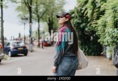 Jeune femme marche dans une rue bordée d'arbres à Amsterdam portant des vêtements décontractés et une casquette de baseball avec des fleurs rouges Banque D'Images