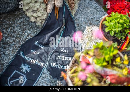 Gros plan d'un ruban commémoratif Waffen SS au cimetière militaire allemand de Normandie, en France, entouré de fleurs vibrantes et d'objets en pierre. Banque D'Images