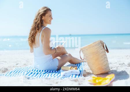 Femme élégante souriante sur la rive de l'océan avec sac de paille et serviette rayée en utilisant des applications sur tablette PC. Banque D'Images