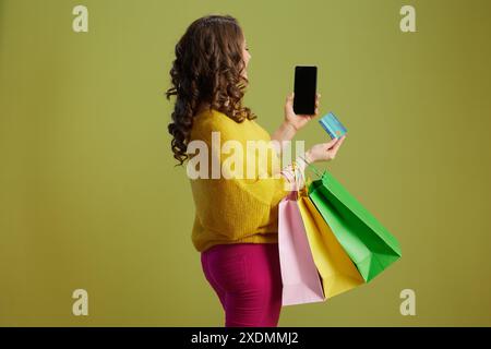 Vu de derrière femme à la mode sur fond vert en pull jaune avec carte de crédit et sacs à provisions montrant l'écran blanc de smartphone. Banque D'Images